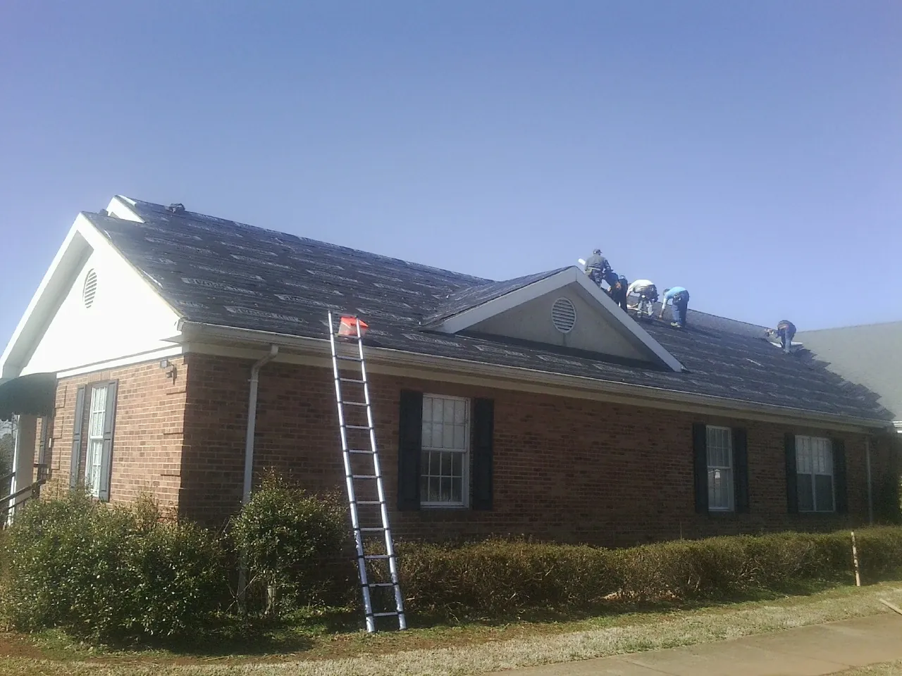 Crew tearing shingles off a brick ranch roof with a ladder set — Metal Roof Repair work by our crew in Jones